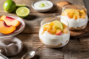 Coppa fredda di ricotta e pesche al lime decorata con menta fresca e granella di pistacchio, servita in una coppetta di vetro su sfondo chiaro